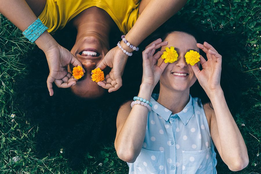 A topdown photograph of two women lying on the grass, holding bright yellow flowers over their faces. 