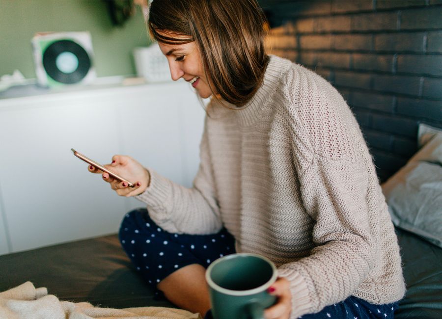 A woman sat on her bed looking through her smartphone with a cup of coffee in her other hand. 