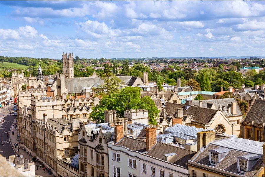 A photo of Oxford’s rooftops and the belltower of the city’s cathedral, with trees and hills in the distance.